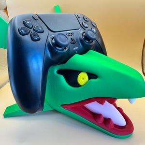 May include: A black PlayStation 5 controller sits on a green, red, and white 3D-printed dragon-themed stand. The stand has a dragon head design with an open mouth, yellow eyes, and a white tongue. The controller is positioned on the stand.
