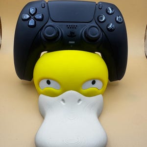 May include: A black PlayStation 5 controller sits above a yellow and white Psyduck-shaped controller holder. The controller holder has a yellow head, white beak, and black eyes. The background is a solid tan colour.