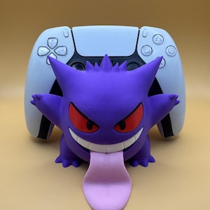 May include: A purple Gengar figure with red eyes and a pink tongue, in front of a white and black game controller. The Gengar figure has a wide grin and spiky features, creating a playful and unique design.
