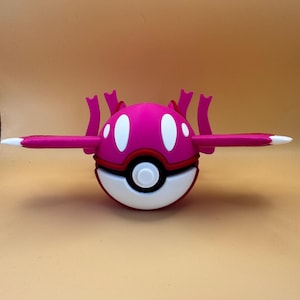 May include: A pink and white toy resembling a Pokémon character. The toy has a spherical body with a red band, white accents, and two horizontal pink arms with white tips. It also features several pink protrusions.