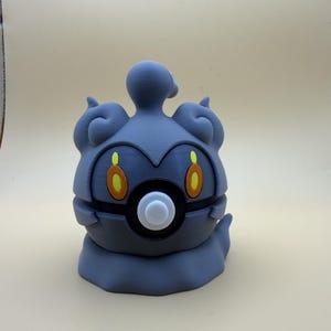 May include: A grey, stylised creature figurine with a rounded, smooth body, featuring two prominent orange and yellow oval-shaped eyes and a central white circular button-like detail on its front, topped with a small, curved protrusion.