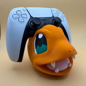 May include: A white and black video game controller resting on an orange, lizard-like character head stand. The character has large green eyes, a light pink mouth, and white fangs. The controller features standard buttons, joysticks, and a central logo.