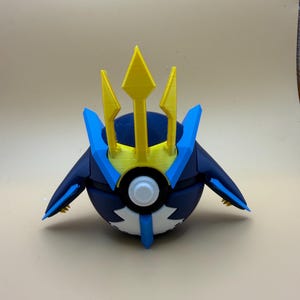 May include: A spherical container featuring a dark blue and white design with a prominent yellow trident-shaped crest on top. The object includes light blue accents, a central white circular button, and two wing-like side extensions with yellow details.