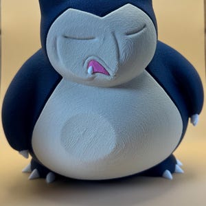 May include: A 3D printed figurine of the character Snorlax, featuring a dark blue body with a cream-coloured belly and face. The figure has closed eyes, a small pink mouth with a single white tooth, and small white claws on its hands and feet.