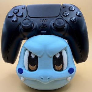 May include: A black PlayStation 5 wireless controller resting on a light blue 3D-printed character stand. The stand features a stylised head with large eyes, dark pupils, small white fangs, and blue circular cheek markings on a smooth surface.