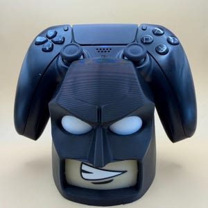 May include: A black video game controller resting on a black 3D-printed stand shaped like a stylised head with a mask. The stand features white eye cutouts and a visible mouth area with a white toothy grin, providing a stable base for the controller.