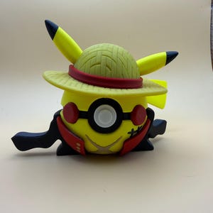 May include: A yellow spherical figure wearing a straw-textured hat with a red band. The figure features two pointed yellow ears with black tips, a central circular eye, two red circular accents, and a red collar-like piece with a stitched design.