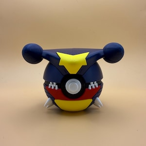 May include: A spherical object featuring a dark blue exterior with a yellow star-shaped emblem on top and two rounded side protrusions. The front displays a central white button, a red band, and white pointed teeth-like accents over a yellow base.