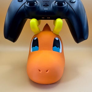 May include: A black PlayStation 5 controller sits above an orange and yellow 3D-printed Charmander figure. The figure has blue eyes and a white outline. The controller is black with multiple buttons and two joysticks.