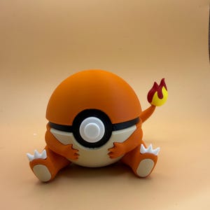 May include: A spherical orange and cream-coloured figurine resembling a creature with a round body, small orange arms, and legs with white claws. The centre features a black and white circular design, and a flame-tipped tail extends from the back.