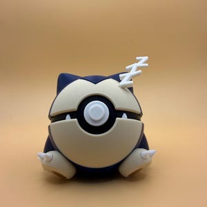 May include: A dark blue and cream-coloured spherical figurine resembling a creature with a central circular button, two small white teeth, and two white Zs attached to the top, representing a sleeping character in a ball-like shape.