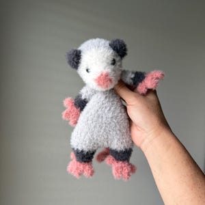 May include: A gray and white crocheted stuffed animal with pink feet and a pink nose. The animal is being held by a hand.