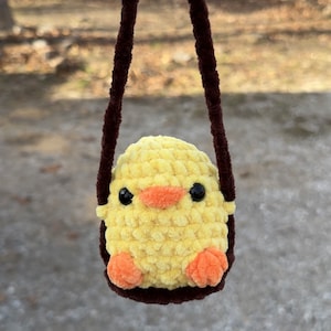 May include: A yellow crocheted chick with orange feet and black eyes is hanging from a brown yarn swing.