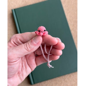 Könnte beinhalten: Ein kleiner rosa gehäkelter Oktopus mit schwarzen Augen und einem lächelnden Mund hängt an einer rosa Schnur. Der Oktopus ist ein kleiner, handgemachter Anhänger.