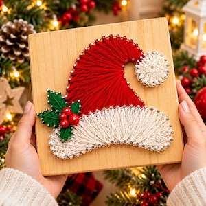 May include: A square wooden plaque featuring a string art design of a Santa hat. The hat is created with red string for the main body, white string for the trim and pom-pom, and green string for holly leaves with three red beads as berries.