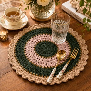 May include: A handmade crochet placemat in shades of green, pink, and tan, with a scalloped edge. A gold spoon and fork with floral patterned handles rest on the placemat, next to a clear glass. A teacup and candle are also visible.