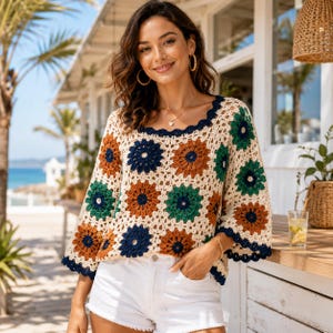 May include: A long-sleeve crochet top featuring a square neckline and a repeating floral pattern in cream, dark blue, rust orange, and forest green. The garment has a scalloped dark blue trim along the neckline and cuffs, with a loose fit.