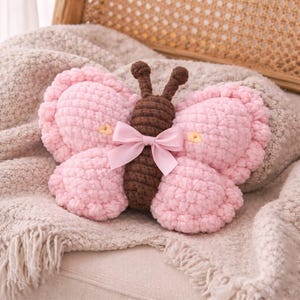 May include: A handmade, plush butterfly pillow in soft pink with a brown body and antennae. A delicate pink bow adorns the center. The pillow is resting on a textured, light-colored blanket, suggesting a cozy, decorative item.