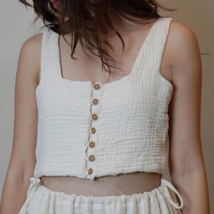 May include: A cream-coloured sleeveless crop top with a square neckline and a front button-down closure featuring seven small brown buttons. The fabric has a textured, crinkled appearance. The word OLIVEE is printed in white at the bottom centre.