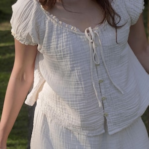 May include: A white textured muslin blouse featuring short puff sleeves with ruffled edges, a scoop neckline, and a front button placket with a matching drawstring tie closure. The fabric has a crinkled appearance and a soft, lightweight texture.