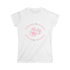 May include: A white short-sleeved t-shirt featuring a circular graphic in pink and green. The design includes line art of three gift boxes and the text "MANIFESTING PR PACKAGES" written in a circular pattern around the central box illustrations.