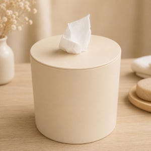 May include: A cylindrical tissue box in a soft beige color. A white tissue is pulled from the top. The tissue box has a simple, modern design, suitable for a bathroom or bedroom. The box is made of a solid material.