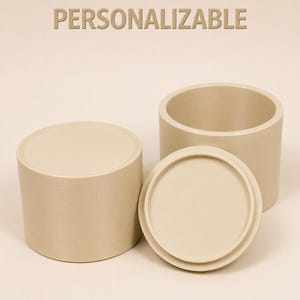 Può includere: Due contenitori cilindrici color beige con coperchi abbinati. Un contenitore è chiuso, mentre l'altro è aperto con il coperchio posizionato davanti. Il testo "PERSONALIZABLE" è stampato in beige sopra. Gli oggetti hanno una finitura liscia.