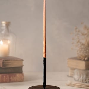 May include: A slender wand featuring a light brown, textured wooden upper section and a smooth, glossy black lower handle. The wand is mounted vertically in a circular, dark brown wooden base, creating a balanced and upright display piece.