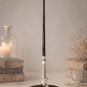 May include: A slender black wand with a metallic silver handle featuring a textured, beaded surface and a crescent moon shape at the base. The wand is displayed vertically, resting on a circular dark brown wooden stand, showcasing a small green gem.