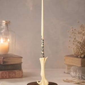 May include: A cream-coloured wand with a textured, organic-shaped handle and a slender, tapered shaft featuring black, elongated markings near the grip. The wand is mounted vertically on a circular, dark brown wooden base.