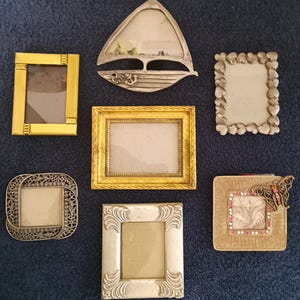 May include: A collection of seven small picture frames in various shapes and finishes. Included are a gold rectangular frame, a silver sailboat-shaped frame, a silver heart-bordered frame, a gold textured frame, a silver filigree frame, a silver ornate frame, and a gold square frame with a butterfly accent.
