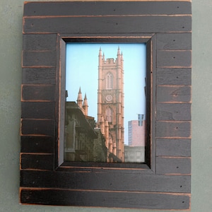 May include: A framed print featuring a tall, ornate building with a clock tower, set against a blue sky. The frame is made of dark, weathered wood, adding a rustic touch to the artwork. The frame measures approximately 12 inches by 10 inches.