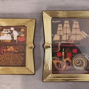 May include: Two rectangular picture frames with a polished gold finish and tiered, stepped edges. The left frame contains a print of figures and text. The right frame displays a three-dimensional scene featuring a sailing ship, red flowers, and a shell.