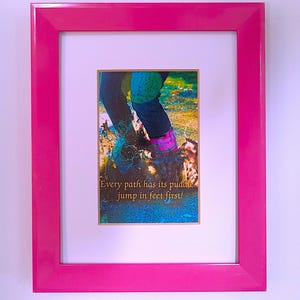 May include: A bright pink rectangular picture frame containing a print of legs in dark pants and pink boots splashing in a puddle. The text on the print reads, Every path has its puddle... jump in feet first! The print has a white mat border.