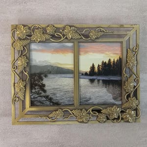 May include: A rectangular double picture frame made of gold-toned metal featuring an ornate relief pattern of grape leaves and clusters of grapes along the border. The frame holds two separate landscape photographs of a lake at sunset with trees.