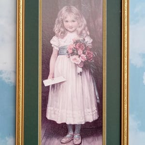 May include: A framed print featuring a young person with long blonde hair wearing a white dress with a blue sash, holding a bouquet of pink and red roses and a small white envelope, standing on a wooden floor in a gold-toned frame.