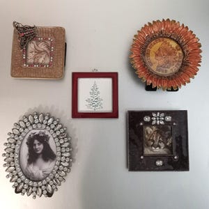 May include: A collection of five decorative frames: a square tan frame with a butterfly and pink crystals, a round orange sunflower frame with a 1902 coin, a small square red frame with a tree illustration, an oval crystal frame, and a dark square frame.