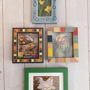 May include: A collection of four rectangular picture frames. The top frame is light blue ceramic with swirl patterns. The middle left frame has a multi-colored striped border. The middle right frame has a wood-grain striped border. The bottom frame is solid green.
