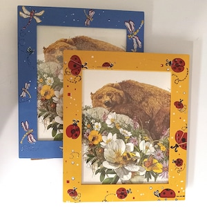 May include: Two rectangular picture frames, one blue and one yellow, each containing an illustration of a brown bear resting on a rock surrounded by white and yellow flowers. The blue frame features dragonfly motifs, while the yellow frame features ladybugs.