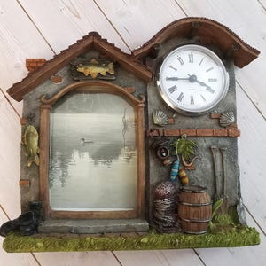 May include: A wall clock shaped like a house with a central picture frame featuring a lake scene. The clock face has Roman numerals. Decorative elements include a fish, a barrel, buoys, and a lantern. The structure has a gray stone texture with brown roof accents and a green base.