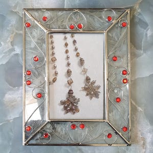 May include: A rectangular glass picture frame with a silver metal border featuring decorative wire swirls and embedded red circular stones. Inside the frame, two beaded necklaces with metallic and brown beads hang against a white background.