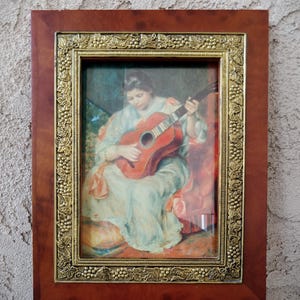 May include: A rectangular wall art piece featuring a print of a person playing a guitar, encased in a frame with a smooth dark wood outer border and an inner gold-toned metal border decorated with a raised relief pattern of grapevines and leaves.