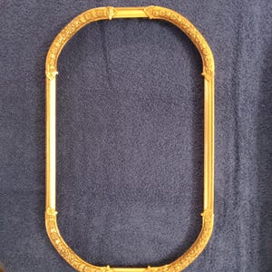 May include: A gold-colored, oval picture frame with ornate floral detailing. The frame has a cream-colored interior and is set against a blue background. The frame's design is symmetrical, with decorative elements at the corners and along the curved edges.