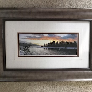 May include: Framed landscape art print featuring a serene lake scene at sunset. The artwork showcases a vibrant sky with orange and purple hues, a dark treeline, and a calm lake. The print is double-matted and encased in a dark brown wooden frame.