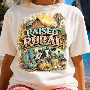 May include: A cream-colored t-shirt featuring a graphic print of a farm scene with a barn, windmill, tractor, sunflowers, a cow wearing a blue bow, and turquoise cowboy boots. The text "RAISED RURAL" is printed in large, stylized lettering.