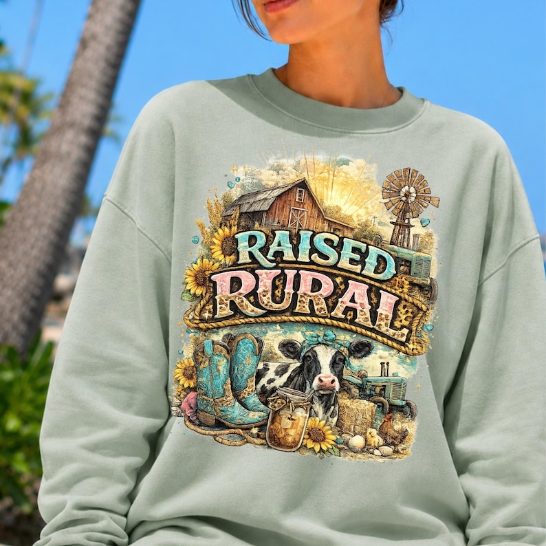 May include: A sage green crewneck sweatshirt featuring a graphic print of a barn, a windmill, a cow, sunflowers, and a pair of turquoise cowboy boots. The text "RAISED RURAL" is printed in a bold, stylized font across the center of the design.