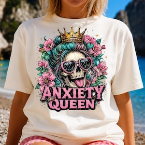 May include: An off-white t-shirt featuring a graphic of a skull wearing a gold crown and pink heart-shaped sunglasses. The skull is surrounded by pink flowers and teal hair, with the text ANXIETY QUEEN printed in bold pink letters below the design.