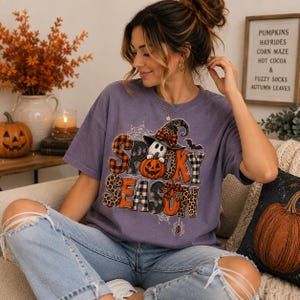 May include: A purple short-sleeve t-shirt featuring a graphic print with the text "SPOOKY SEASON" in various patterns including plaid, leopard print, and orange. The design includes a ghost wearing a witch hat, a pumpkin, and spiderwebs.