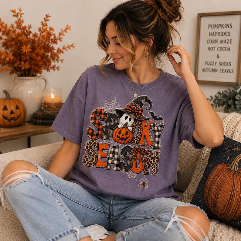 May include: A purple short-sleeve t-shirt featuring a graphic print with the text "SPOOKY SEASON" in various patterns including plaid, leopard print, and orange. The design includes a ghost wearing a witch hat, a pumpkin, and spiderwebs.