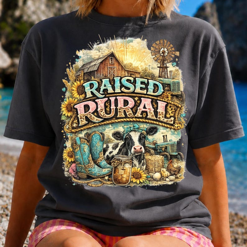 May include: A charcoal gray t-shirt featuring a graphic print with the text RAISED RURAL. The design includes a barn, a windmill, a cow with a blue bandana, turquoise cowboy boots, a mason jar, sunflowers, and a tractor in a farm setting.